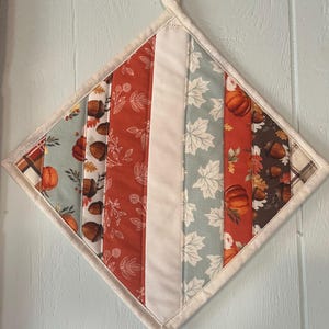 May include: A diamond-shaped quilted potholder featuring vertical fabric strips with autumn-themed patterns including pumpkins, acorns, and maple leaves in shades of orange, brown, cream, and sage green, finished with a cream-colored fabric loop.