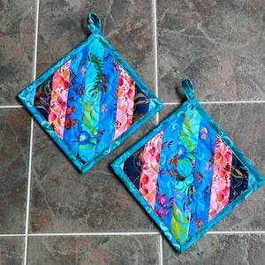 May include: Two square quilted pot holders featuring a diagonal striped pattern in shades of blue, pink, orange, and green. The fabric displays a colorful aquatic print with fish and seaweed motifs, finished with a teal border and a hanging loop.