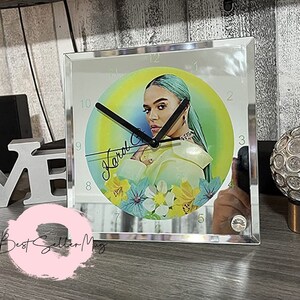 May include: A square mirrored clock with a round face featuring a colorful portrait of a woman with blue hair. The clock face has a rainbow gradient background with yellow flowers and the woman's signature "Kard" in the center.