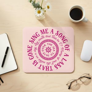 May include: A pink square mousepad with the text "SING ME A SONG OF A LASS THAT IS GONE" in a circular design. The design includes a ship's wheel and decorative knotwork. A cup of coffee, a mouse, and glasses are on the wooden desk.