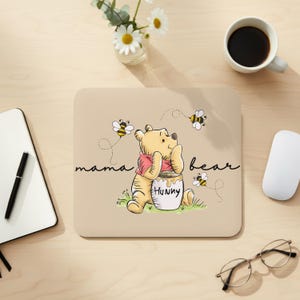 Puede incluir: Alfombrilla de ratón beige con un oso de dibujos animados, un tarro de miel y abejas. El texto "mama bear" está escrito en cursiva. El oso lleva una camisa rosa. También se ven un ratón de ordenador, una taza de café y gafas.