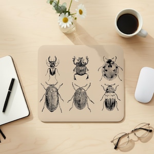 Pode incluir: Tapete de rato bege com seis ilustrações detalhadas a preto e branco de várias espécies de besouros. O tapete do rato é retangular com cantos arredondados. Um bloco de notas, caneta, chávena de café e óculos também são visíveis.