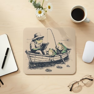 May include: Beige mousepad featuring a whimsical illustration of three frogs in a rowboat. One frog reads a book, another fishes, and the third looks on. The frogs wear hats and glasses. The design is in shades of green, brown, and beige.