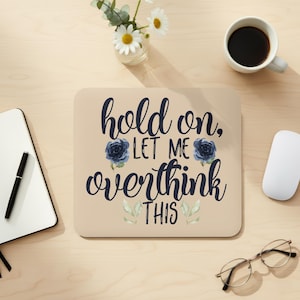 May include: A beige mousepad with the text "hold on, LET ME overthink THIS" in dark blue script. The design includes dark blue rose illustrations and green leaves. A white mouse, a cup of coffee, and a notebook are also visible.