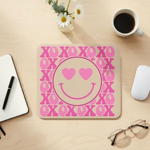 Puede incluir: Una alfombrilla de ratón cuadrada beige con una carita sonriente rosa con ojos de corazón en el centro. La alfombrilla está rodeada por las palabras "XOXO" en rosa. Un ratón de ordenador blanco, una taza de café y un cuaderno están sobre el escritorio de madera.
