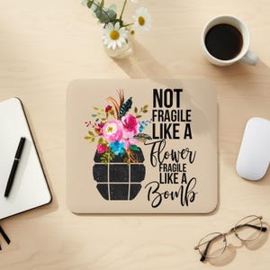 May include: A beige mousepad with the text "NOT FRAGILE LIKE A FLOWER FRAGILE LIKE A BOMB." The mousepad features a floral design with pink, blue, and red flowers. A black coffee mug, a white mouse, and a pair of glasses are also visible.
