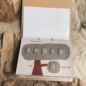 May include: A brown and beige illustrated book with a tree and the name "ANDIE" spelled out with letter tiles. The book also says "My name is" and "I am 3 years old".