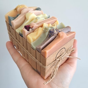 May include: A stack of six handmade soap bars wrapped in brown paper and twine. The soaps are various colours, including pink, green, yellow, and grey. The soaps are decorated with dried flowers and herbs.