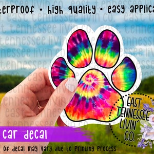 May include: A colorful tie-dye paw print sticker with a black outline. The sticker is on a white background and is being held by a hand. The sticker says "East Tennessee Livin' Co."