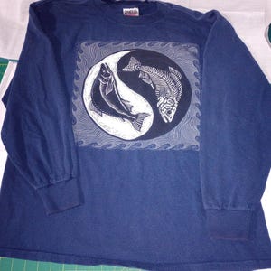May include: Navy blue long-sleeve shirt featuring a Yin Yang design with a fish and a fish skeleton. The graphic is in black and white, with a wave pattern border. The shirt has a crew neck and ribbed cuffs.