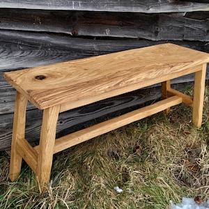 Op de afbeelding: Een rechthoekige houten bank gemaakt van lichtbruin hout met een zichtbaar natuurlijk houtnerfpatroon. De bank heeft een glad, plat bovenoppervlak met een opvallende donkere knoest en vier stevige, schuine poten verbonden door een horizontale steun.