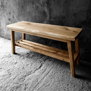 May include: A rectangular wooden bench featuring a natural light brown wood grain finish, a smooth flat top surface, and a lower slatted shelf supported by four sturdy legs. The bench is constructed with visible wood knots and organic textures.