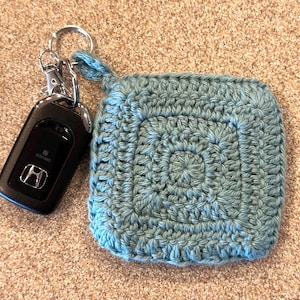 Op de afbeelding: Een gehaakte, vierkante sleuteletui in een zachte teal kleur, met een lus om aan een sleutelhanger te bevestigen. Een zwarte autosleutel met een zilveren sleutelhanger ligt naast de etui. De etui is handgemaakt en heeft een getextureerde uitstraling.