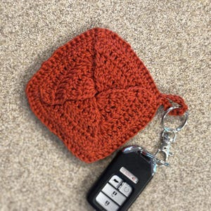 Op de afbeelding: Een roestkleurige, gehaakte sleutelhangerhouder met een decoratief patroon. De houder is bevestigd aan een zilveren sleutelring en een zwarte autosleutelhanger. De sleutelhangerhouder is vierkant.