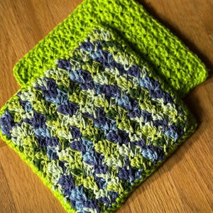 Könnte beinhalten: Zwei handgefertigte gehäkelte Waschlappen. Einer ist einfarbig hellgrün, der andere ist eine Mischung aus Grün, Blau und Lila. Die Waschlappen sind quadratisch und haben ein strukturiertes Stichmuster. Sie liegen auf einer Holzoberfläche.