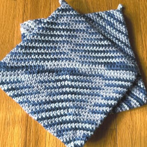 Può includere: Due presine quadrate fatte a mano, nei toni del blu e del bianco. Le presine sono lavorate all'uncinetto con un motivo a righe e hanno un piccolo anello in un angolo per appenderle. Le presine sono appoggiate su una superficie di legno.