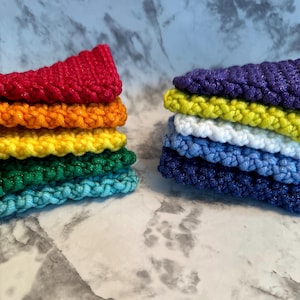 May include: Two stacks of crocheted items in a rainbow of colors. One stack features red, orange, yellow, green, and turquoise. The other stack has purple, yellow, white, light blue, and navy blue. The items are made of a textured yarn.