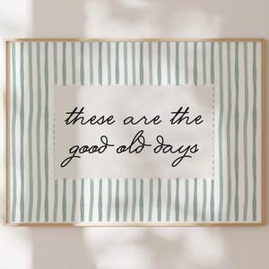 May include: A framed print with a light green and white striped background. The center features a white rectangle with the handwritten text "these are the good old days" in black script. The frame is a light wood color.