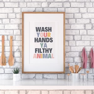 May include: A framed print with the text "WASH YOUR HANDS YA FILTHY ANIMAL" in colorful letters on a white background. The frame is made of wood and has a light brown finish.