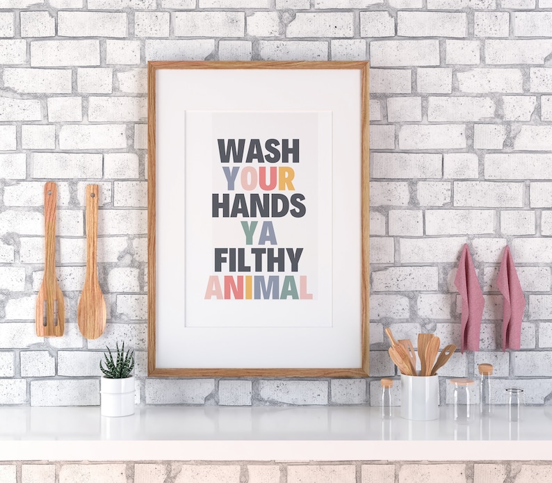 May include: A framed print with the text "WASH YOUR HANDS YA FILTHY ANIMAL" in colorful letters on a white background. The frame is made of wood and has a light brown finish.