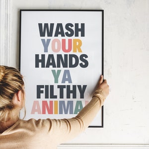 May include: A framed poster with the text "WASH YOUR HANDS YA FILTHY ANIMAL" in colorful letters. The poster is being hung on a white wall.