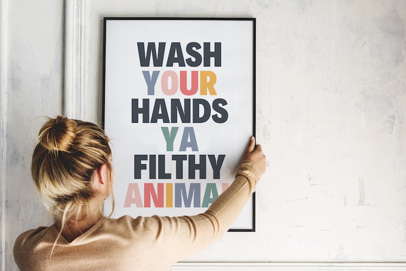 May include: A framed poster with the text "WASH YOUR HANDS YA FILTHY ANIMAL" in colorful letters. The poster is being hung on a white wall.