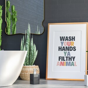 May include: A gold framed print with the text "Wash Your Hands Ya Filthy Animal" in colorful letters. The print is hanging on a black brick wall in a bathroom setting. There are two plants, a white bathtub, and two soap dispensers in the image.