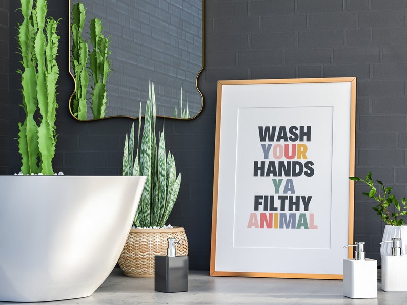 May include: A gold framed print with the text "Wash Your Hands Ya Filthy Animal" in colorful letters. The print is hanging on a black brick wall in a bathroom setting. There are two plants, a white bathtub, and two soap dispensers in the image.