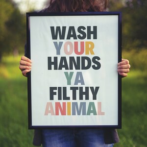 May include: A framed poster with the text "WASH YOUR HANDS YA FILTHY ANIMAL" in colorful letters. The poster is being held by a person in a green field.