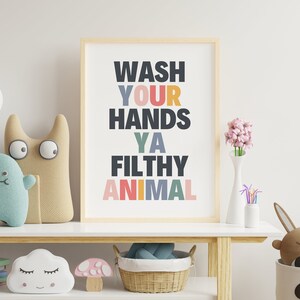 May include: A framed print with the text "Wash Your Hands Ya Filthy Animal" in a colorful, playful font. The print is hanging on a white wall above a wooden shelf with various toys and decor.