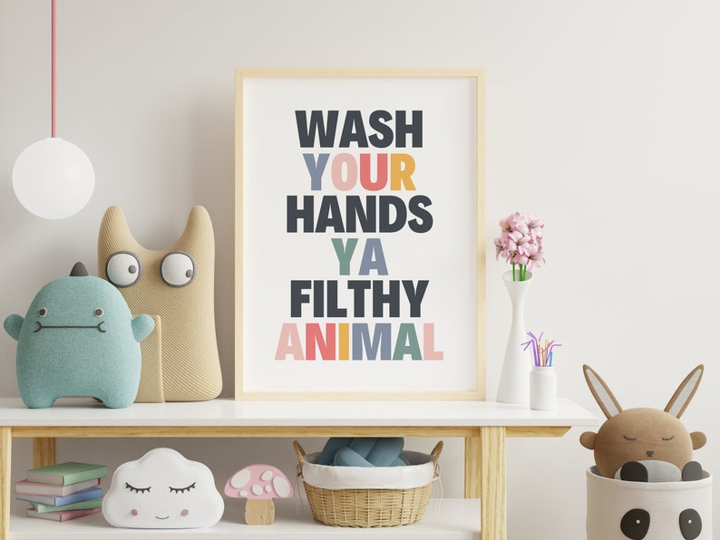 May include: A framed print with the text "Wash Your Hands Ya Filthy Animal" in a colorful, playful font. The print is hanging on a white wall above a wooden shelf with various toys and decor.