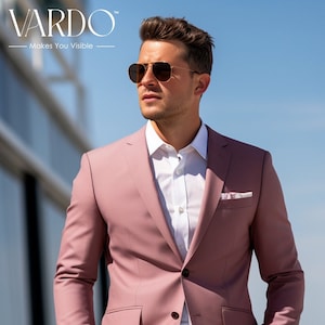 May include: A dusty rose suit jacket and matching trousers worn over a crisp white button-down shirt. The jacket features two dark buttons, flap pockets, and a pocket square. The ensemble is paired with gold-rimmed sunglasses and a brown belt.
