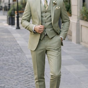 K&ouml;nnte beinhalten: Ein dreiteiliger salbeigr&uuml;ner Anzug mit einem wei&szlig;en Hemd und einer gemusterten Krawatte. Der Anzug besteht aus Jacke, Weste und Hose. Braune Lederschuhe vervollst&auml;ndigen das Outfit. Ein Knopflochblume ist am Revers der Jacke befestigt.