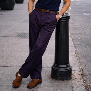 Puede incluir: Una persona con una camisa azul marino, pantalones plisados ​​de color púrpura oscuro, un cinturón de cuero marrón y mocasines de gamuza marrones. Se ve un reloj de pulsera. La persona está apoyada en un poste negro en una calle.