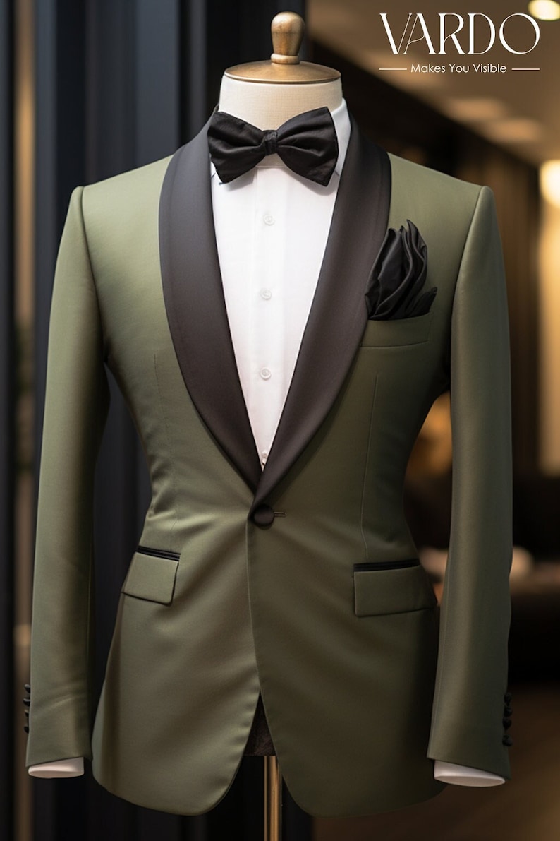 May include: An olive green tuxedo jacket featuring a black satin shawl lapel, a single black button closure, and two flap pockets. The jacket is worn over a white button-down shirt with a black bow tie and a black pocket square. Text reads VARDO.