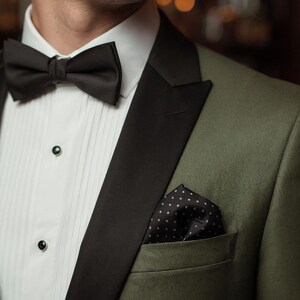 May include: An olive green tuxedo jacket with black lapels, a white pleated dress shirt with black buttons, a black bow tie, and a black pocket square with white polka dots. The text VARDO Makes You Visible is visible in the top right corner.