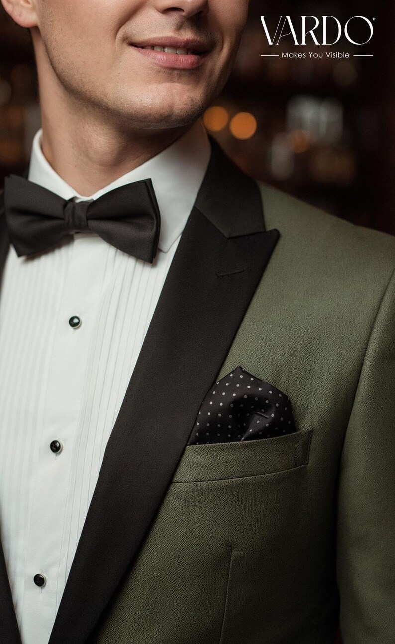 May include: An olive green tuxedo jacket with black lapels, a white pleated dress shirt with black buttons, a black bow tie, and a black pocket square with white polka dots. The text VARDO Makes You Visible is visible in the top right corner.