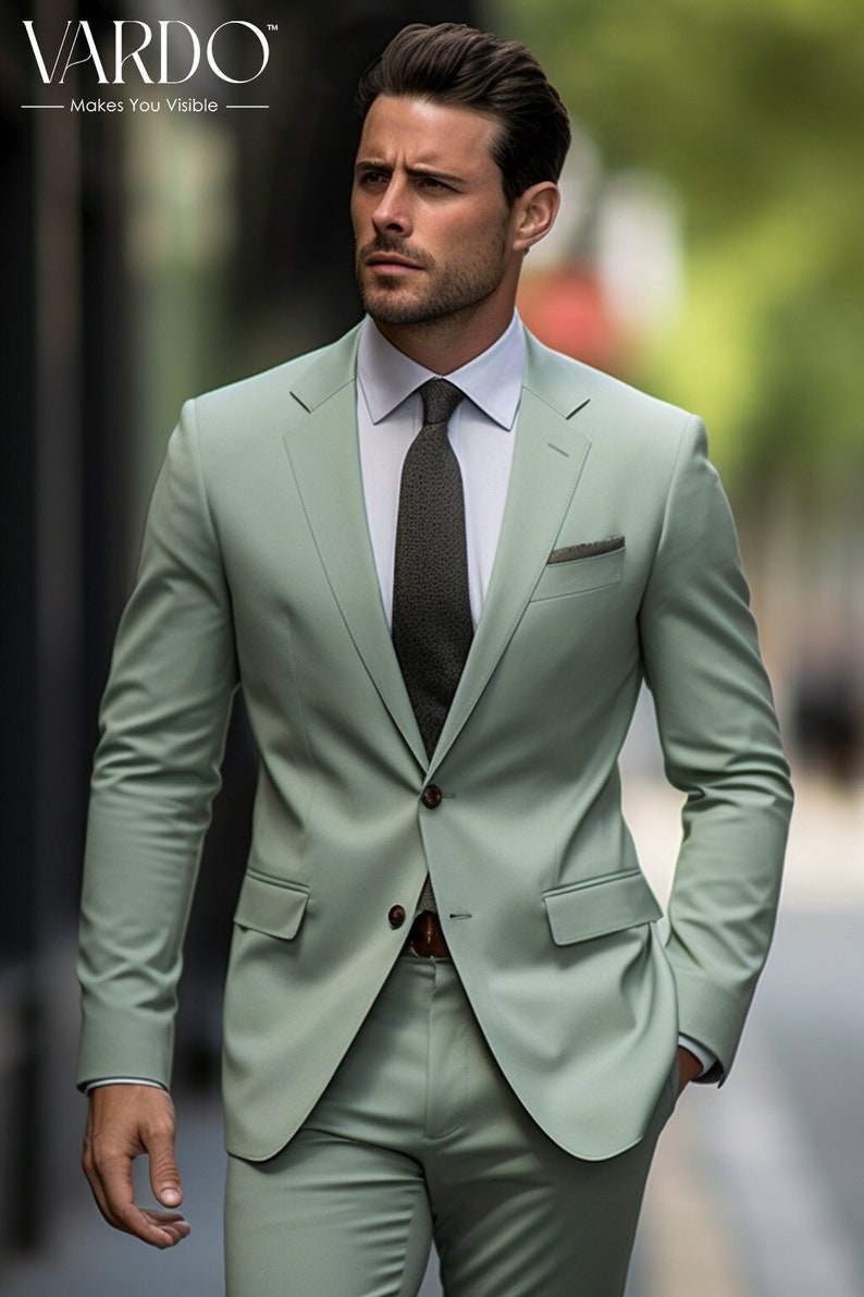 May include: A man wearing a light green suit with a dark green tie. The suit jacket has a single button closure and a pocket square. The man is standing in front of a blurred background.