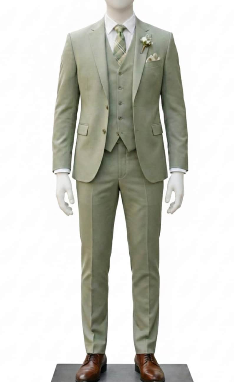 K&ouml;nnte beinhalten: Ein dreiteiliger Anzug in gedecktem Salbeigr&uuml;n, pr&auml;sentiert auf einer Schaufensterpuppe. Der Anzug besteht aus Blazer, Weste und Hose. Ein wei&szlig;es Hemd, eine gemusterte Krawatte und ein Einstecktuch setzen Akzente. Braune Lederschuhe runden das Outfit ab.