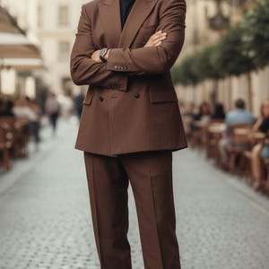 May include: A brown double-breasted suit with matching trousers. The suit features a classic design with a notched lapel and a two-button closure. The person is wearing a black shirt and brown leather shoes. The watch has a silver band.