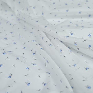 May include: White fabric with a delicate floral pattern. The fabric features small blue flowers and tiny black accents scattered across its surface. The material is soft and flowing, with visible folds and creases.