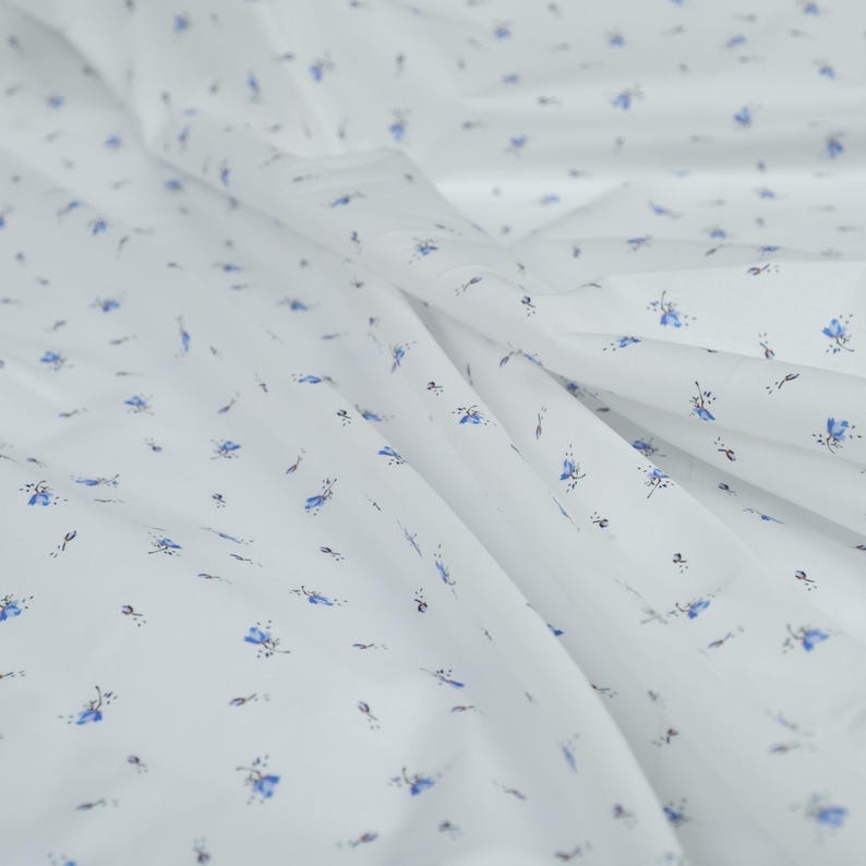 May include: White fabric with a delicate floral pattern. The fabric features small blue flowers and tiny black accents scattered across its surface. The material is soft and flowing, with visible folds and creases.