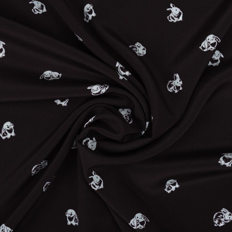 May include: Black fabric with a repeating pattern of white cartoon bunnies in various poses. The fabric is draped, showing folds and shadows, and the bunnies are scattered across the surface.
