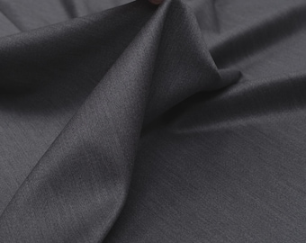 May include: A close-up of a folded piece of gray fabric. The fabric is held up by a hand, revealing its texture and drape. The material appears to be smooth with a subtle sheen, suggesting a high-quality textile.