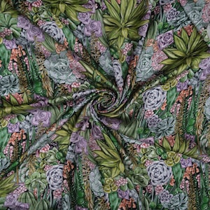 Könnte beinhalten: Ein Stoff mit einem detaillierten botanischen Druck, der verschiedene Sukkulenten in Grün-, Lila- und Orangetönen zeigt. Das Design umfasst Aloe Vera, Echeverien und andere Wüstenpflanzen und erzeugt ein reichhaltiges, strukturiertes Muster. Der Stoff ist gefaltet.