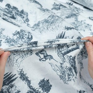 May include: White fabric with a black and white print of a landscape scene. The fabric is being held in a hand, showing the texture of the material.