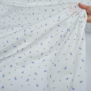 May include: White fabric with a delicate floral pattern. Small blue flowers and tiny black stems are scattered across the material. The fabric is held up, showing its lightweight and flowing texture, suitable for apparel or home decor.