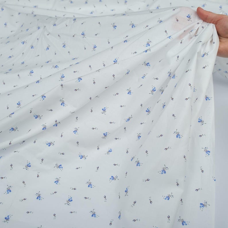 May include: White fabric with a delicate floral pattern. Small blue flowers and tiny black stems are scattered across the material. The fabric is held up, showing its lightweight and flowing texture, suitable for apparel or home decor.