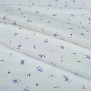 May include: White fabric with a delicate floral pattern. Small blue flowers with dark stems and leaves are scattered across the surface. The fabric is folded, creating soft shadows and highlighting the texture of the material.