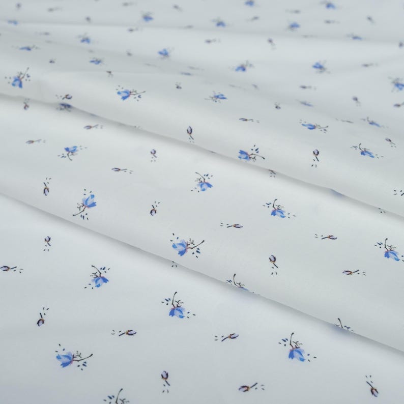 May include: White fabric with a delicate floral pattern. Small blue flowers with dark stems and leaves are scattered across the surface. The fabric is folded, creating soft shadows and highlighting the texture of the material.